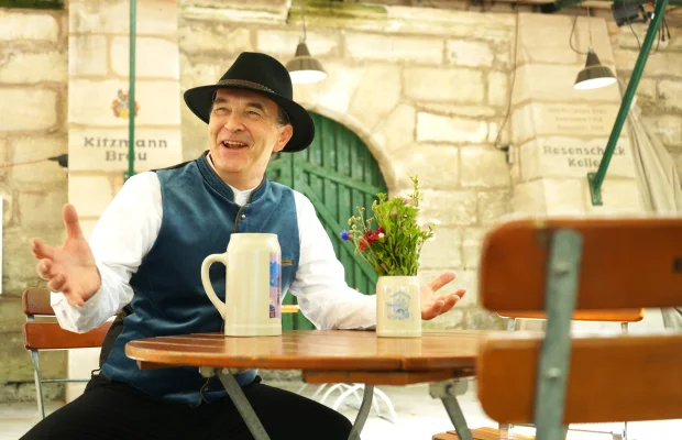 Wir sehen den kostümierten Stadtführer als Bierkutscher Schorsch, sitzend vor dem Kitzmann Keller in Erlangen, stilecht mit einem Ton-Bierkrug.